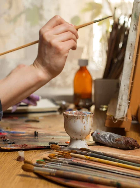 The artist's hands hold a brush near a canvas with a picture in an art studio. The concept of lifestyle and hobby. Close-up, selective focus