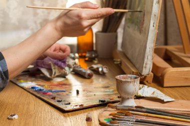 The artist is working on a Still Life. Brush strokes on the canvas. Studio with a desktop easel and brushes on the table. Close-up, Focus on the bunch of brushes in the foreground