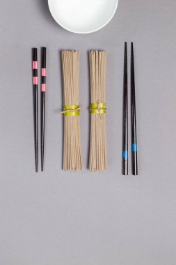 Japon mutfağını karabuğday erişteli soba ile pişirme fikri. İki paket kuru erişte, yemek çubuğu ve gri arka planda boş bir çorba kasesi..