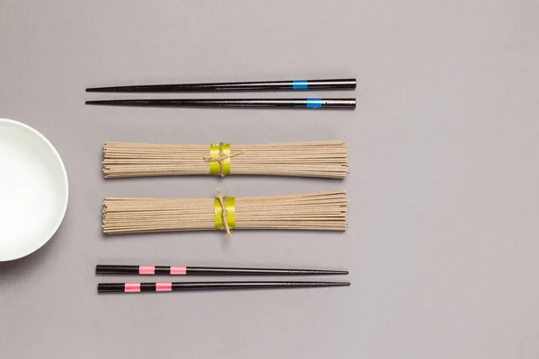 Dos porciones de fideos de soba japoneses de trigo sarraceno seco, dos pares de palillos y un tazón de sopa. Pisos sobre un fondo gris, copia del espacio, horizontal — Foto de Stock