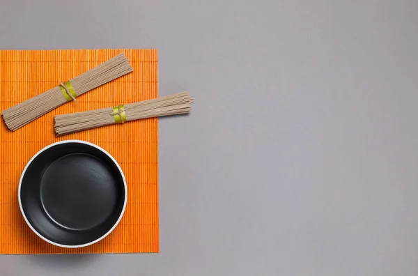 Dos paquetes de fideos secos de trigo sarraceno soba, en una servilleta de bambú naranja, y un bol de sopa negro. Pisos sobre fondo gris, con espacio — Foto de Stock