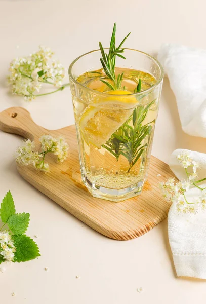 A glass of sparkling elderflower drink with lemon slices and rosemary, on a wooden board. Close-up on a light background,with elderberry flowers and a linen napkin, vertical