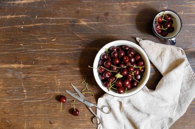 Metal bir kasede kirazlar ve siyah ahşap bir masada eski bir makas ve keten bir havluyla kupalar. Yatay, boşluklu.