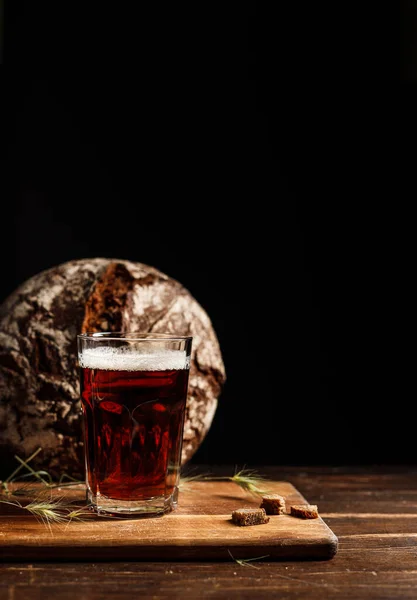 Un verre avec du seigle foncé kvass ou de la bière, avec une mousse épaisse. Sur une planche de bois avec craquelins et épillets. Pain rond de seigle en arrière-plan — Photo