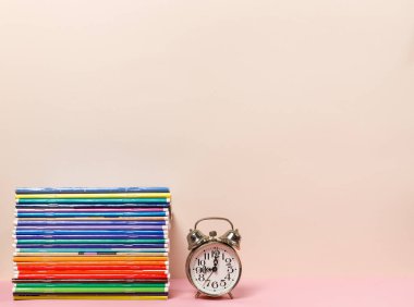 Masanın üzerinde renkli defterler ve çalar saat yığını var. Uzayı olan yatay bir çerçeve. Pastel renkler.