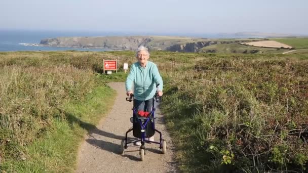 Gepensioneerde m/v actieve oude dame lopen naar camera met drie wielen trolley mobiliteit steun op kust pad door kust scène Bedruthan Noord Cornwall Engeland Uk — Stockvideo