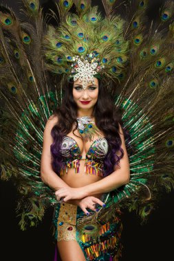 Kafasında tüyleri olan esmer bir kadın. Arka planda gür tavus kuşu tüyleri var..