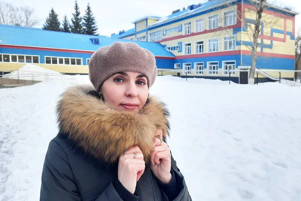 A woman in winter clothes stands in the snow near city school.