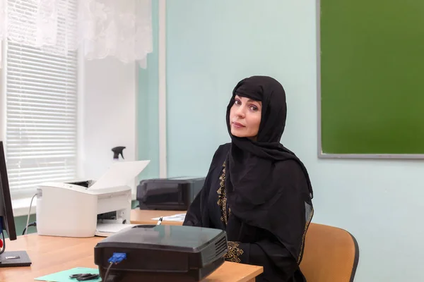 A Muslim teacher in national dress sits at a desk in the office.