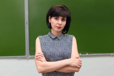 Esmer bir öğretmen kollarını kavuşturmuş tahtada duruyor..