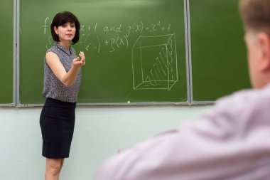 Öğretmen tahtada duruyor ve öğrenciye el hareketiyle cevap vermesini söylüyor..