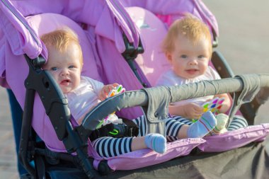 İki ikiz kardeş ellerinde oyuncaklarla bebek arabasında yan yana oturuyorlar..