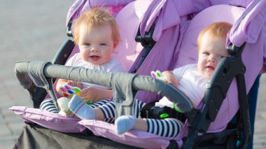 Yazlık hafif elbiseli iki kız kardeş ellerinde parlak oyuncaklarla bebek arabasında oturuyorlar..