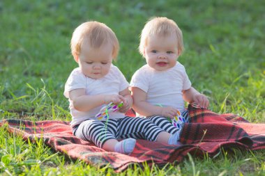 Parkta bir battaniyenin üzerinde birbirine benzeyen iki kız ellerinde parlak oyuncaklarla uzakta şaşkınlık içinde bakıyorlar..