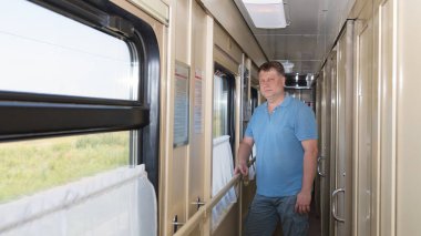 35-40 yaşlarında sarışın bir adam trenin koridorunda dikiliyor ve pencereden dışarı bakıyor..