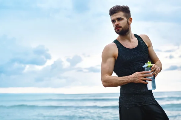 Homem da aptidão com a garrafa de água que descansa após o exercício na praia — Fotografia de Stock
