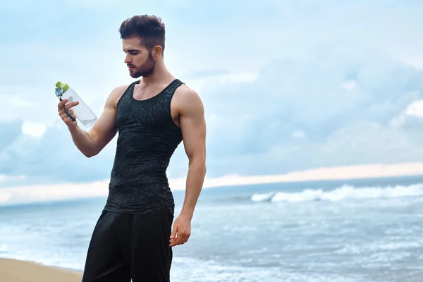 Hombre de fitness con botella de agua Descansando después del entrenamiento en la playa —  Fotos de Stock