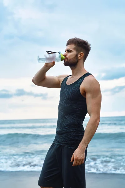 Hombre bebiendo agua refrescante después del entrenamiento en la playa. Bebe. — Foto de Stock