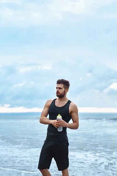 Homme de remise en forme avec bouteille d'eau se reposant après l'entraînement à la plage — Photo