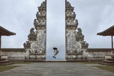 Beautiful Girl Full-Length Portrait Near Gates Of Heaven In Pura Lempuyang Temple In Bali, Indonesia. Happy Young Woman Doing Back Band Jump Near Ancient Architecture In Asia. clipart