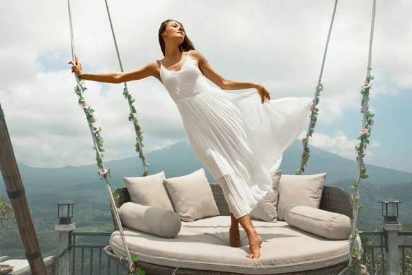 美丽的女孩站在天空摇曳的肖像在巴厘岛，印度尼西亚。穿着白衣的年轻女子随风飘扬，挂着长椅，花朵迎山飘扬. — 图库照片