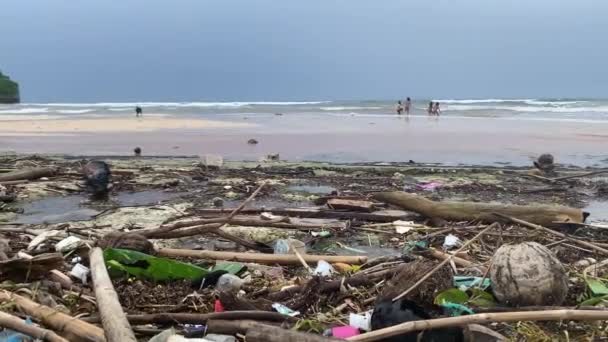 Niños y perros jugando en la playa del océano desperdiciado en Indonesia. Los niños atrapan olas después de la tormenta y se divierten en la costa cubierta con plástico, basura, basura y basura sólida. — Vídeos de Stock