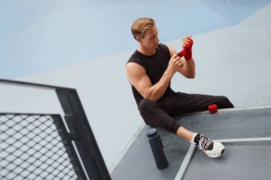 Beton duvara karşı boks bandıyla elini ve bileğini saran genç adam. Yakışıklı, beyaz tenli, kaslı sporcu. Moda dünyasında spor kıyafetleri içinde. Açık havada oturuyor..