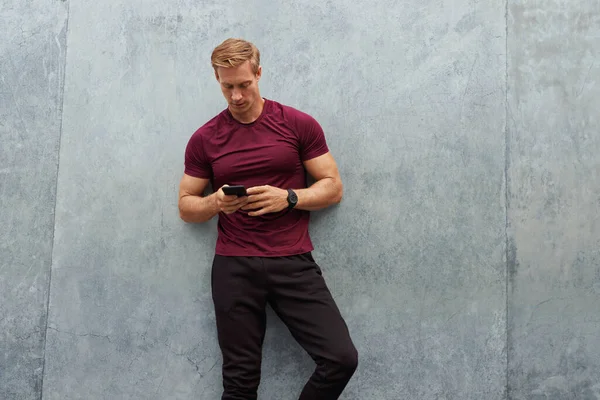 Молода людина в спортивному одязі, використовуючи смартфон проти бетонної стіни. Красивий Кавказький спортсмен з сильним м'язовим тілом позує на відкритому повітрі . — стокове фото