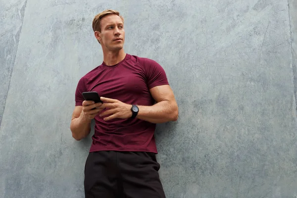 Young Man In Sportswear Using Smartphone Against Concrete Wall. Handsome Caucasian Sportsman With Strong Muscular Body Posing Outdoors.