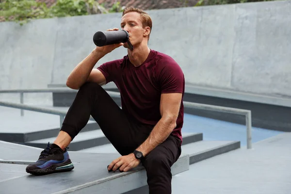 Junger Mann trinkt nach intensivem Training auf Betonstufen aus der Flasche Schöner kaukasischer Sportler mit starkem muskulösem Körper in modischer Sportbekleidung mit Pause. — Stockfoto