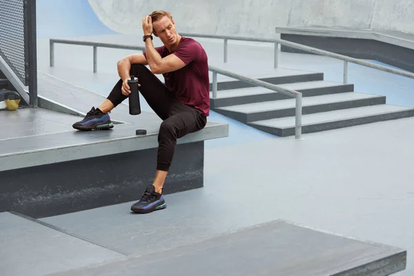 Молодой человек пьет из бутылки после интенсивной тренировки на бетонных ступенях. Красивый кавказский спортсмен с сильным мускулистым телом в моде. — стоковое фото