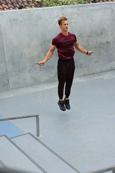 Joven saltando sobre la cuerda contra la pared de hormigón al aire libre. Guapo atleta caucásico con fuerte cuerpo muscular en ropa deportiva de moda calentamiento antes de entrenamiento intenso. —  Fotos de Stock