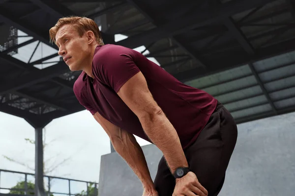 Junger Mann im verschwitzten Hemd ruht sich nach intensivem Training gegen Betonwand aus. Schöner kaukasischer Sportler mit kräftigem muskulösen Körper in modischer Sportbekleidung sieht müde und erschöpft aus. — Stockfoto