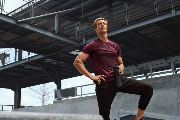 Hombre joven con camisa sudorosa descansando después de un intenso descanso de entrenamiento contra la pared de hormigón. Guapo atleta caucásico con fuerte cuerpo muscular en ropa deportiva de moda se ve confiado. —  Fotos de Stock