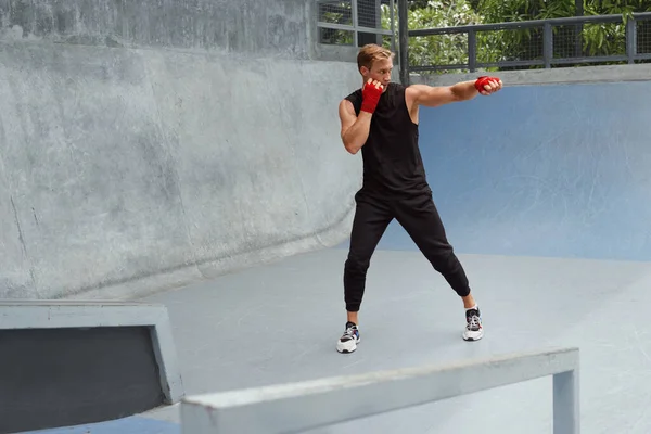 Hombre joven se para en la postura de boxeo y haciendo entrenamiento de perforación. Guapo atleta caucásico con fuerte cuerpo muscular en ropa deportiva de moda y envoltura de mano en las muñecas contra la pared de hormigón. — Foto de Stock