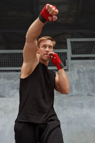 Young Man Stands In Boxing Pose And Doing Punching Workout. Handsome Caucasian Sportsman With Strong Muscular Body In Fashion Sportswear And Hand Wrap On Wrists Against Concrete Wall. — Stock Photo, Image