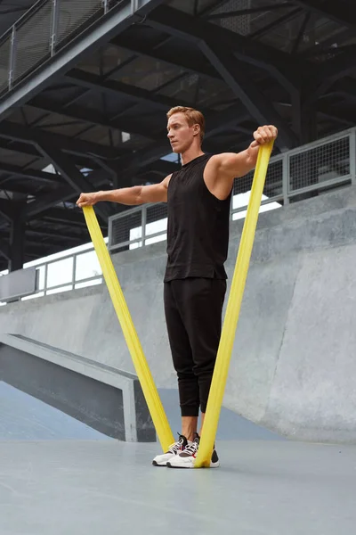 Jovem se alongando com banda de resistência contra parede de concreto ao ar livre. Bonito desportista caucasiano com corpo muscular forte na moda Sportswear fazendo exercícios de alongamento. — Fotografia de Stock