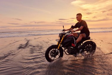 Ocean Beach 'teki Adam ve Motosiklet Güzel Tropikal Gün Batımında. Endonezya, Bali 'de Kum Sahili' nde Motosikletli Yakışıklı Motorcu.