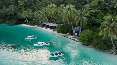 Kaimana Adası, Raja Ampat 'ta Palmiye Ağaçları Arasında Kulübeleri olan Okyanus Kıyısı' na yakın tekneler. Papua, Endonezya 'daki tropik tatil beldesi yakınlarındaki Turkuaz Gölündeki Drone On Water Transport' tan çarpıcı bir görüntü..