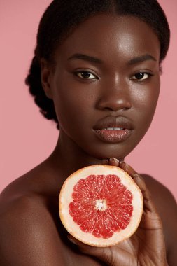 Güzel bir Afrikalı kızın portresi. Elinde bir dilim greyfurt var. Ciddi bir genç kadın kameraya bakıyor. Cilt bakımı. Pembe arka planda izole edilmiş. Stüdyo çekimi