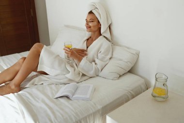 Pleased european girl with closed eyes laying on bed and drink lemonade. Young beautiful woman with under eye patch on face wear bathrobe and wrapped bath towel on head. Concept of rest. Home bedroom clipart