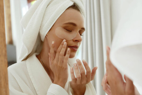 European girl applying under eye patch on face and watch in mirror. Young concentrated beautiful woman with closed eye wear bathrobe and wrapped bath towel on head. Concept of face skin care