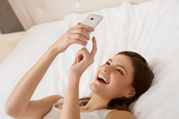 Retrato de una mujer feliz acostada en la cama usando un teléfono inteligente — Foto de Stock