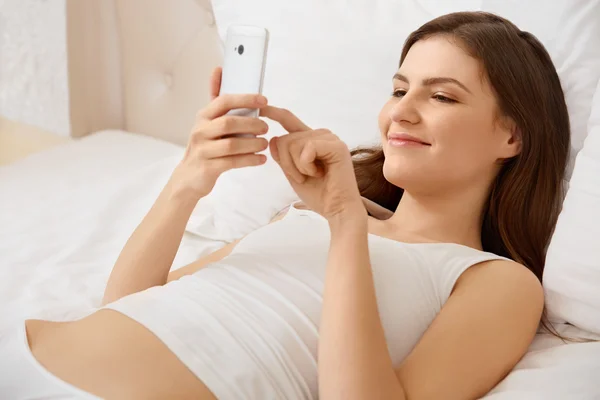 Retrato de una mujer feliz acostada en la cama usando un teléfono inteligente — Foto de Stock