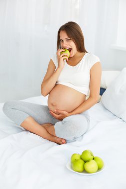 Pregnant Young Woman holding Apple while sitting on the Bed. Hea