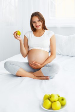 Pregnant Young Woman holding Apple while sitting on the Bed. Hea