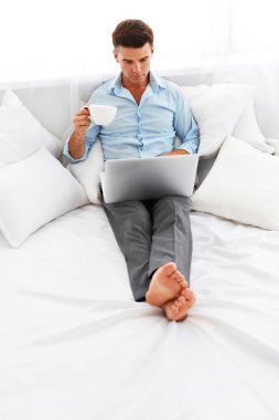 Man lying in bed with his notebook, drinking coffee.