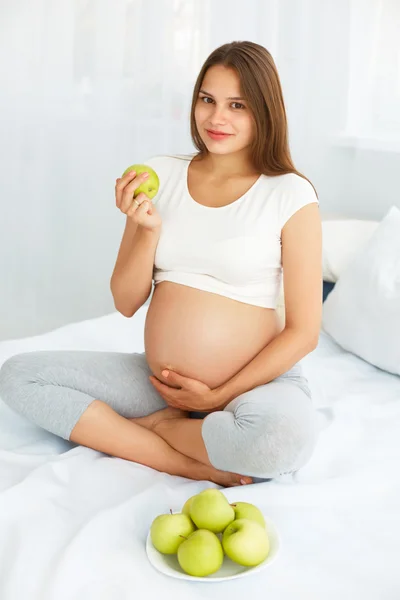 Giovane donna incinta che tiene Apple mentre è seduta sul letto. Hea. — Foto Stock