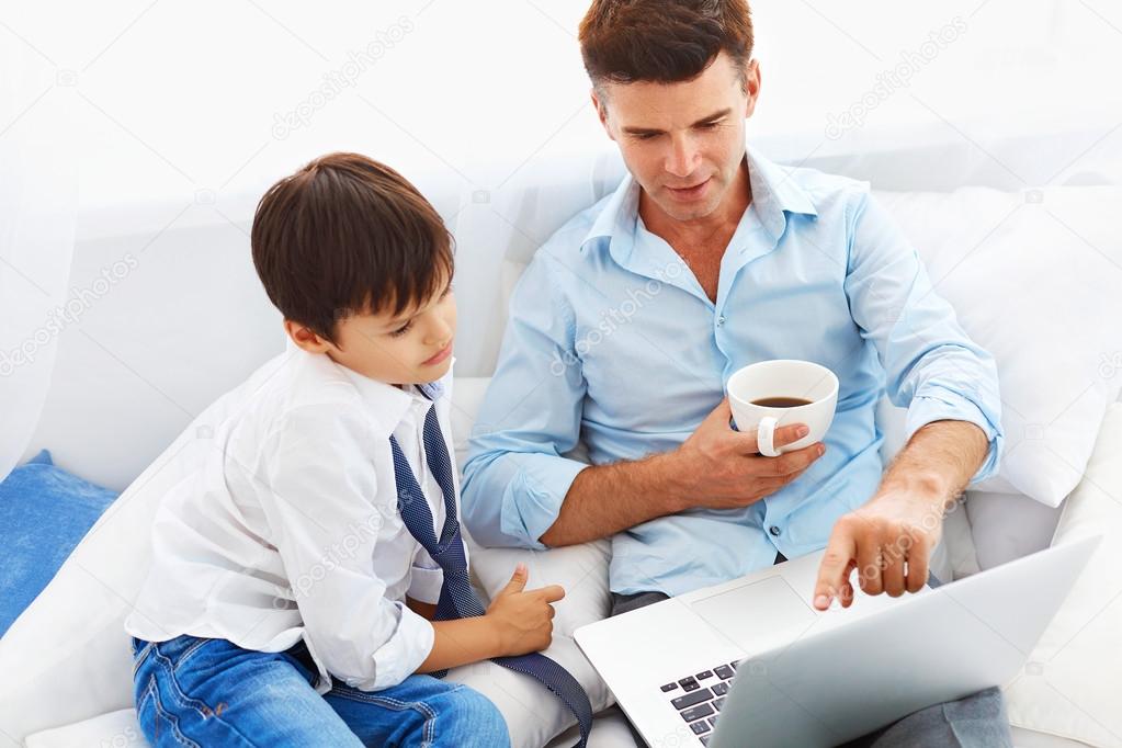 Father drinking coffee and teaching son how to use notebook.