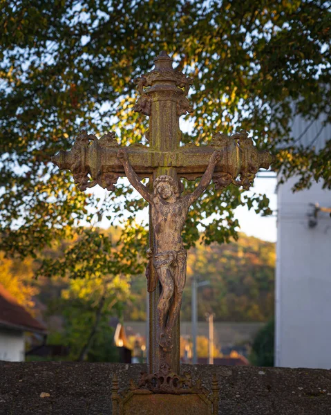 Jesus on the cross - antique iron cross with figure of Christ. Crucifix at cemetery grave, trees and colorful leaves in background